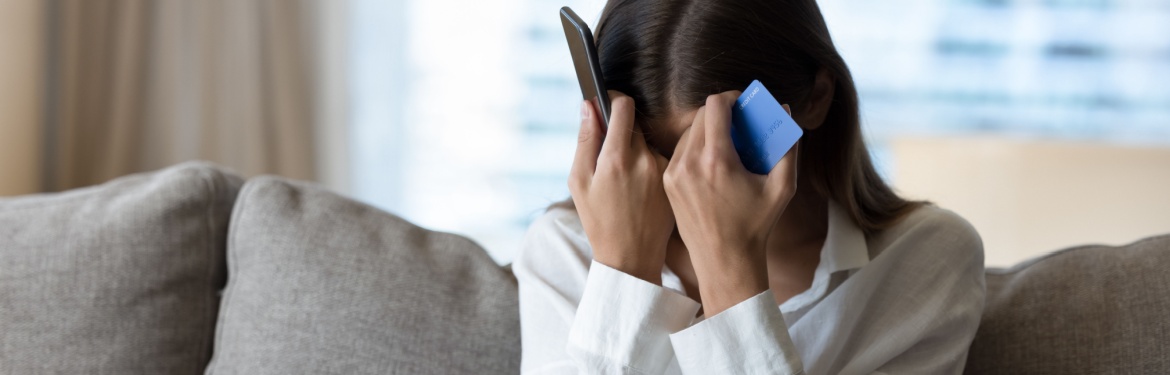 A woman holds her head with a phone in one hand and a bank card in the other.